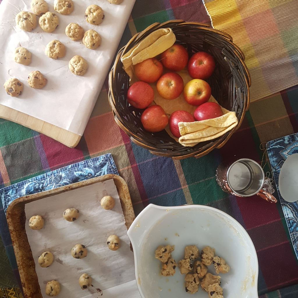 Chocolate Chip & Chia Seed Two-Bite Cookies - Table top view of cookie dough balls in a bowl and lined on a tray. A basket of apples and a mug also sit on the table top